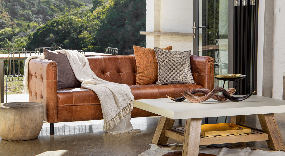 A nature background with a buttoned leather couch styled with three brown-grey cushions and a cream throw together with a white and wooden coffee table styled with animal horns